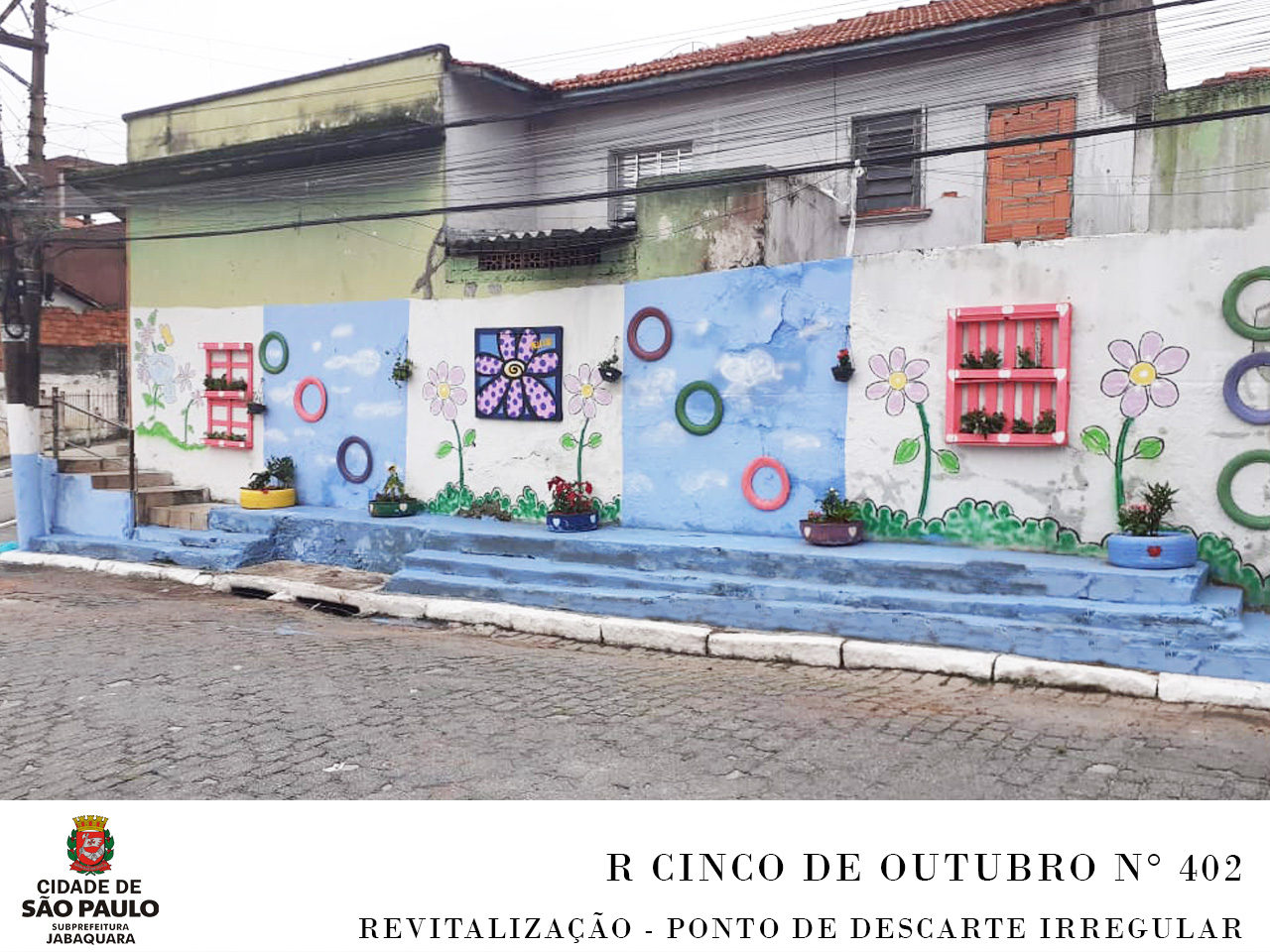 Local revitalizado pela Subprefeitura, contendo as cores azul e branco na parede, desenho de flores nas cores roxo e lilás, o local é um muro na na esquina da rua com 3 degraus. 